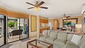 Sala de estar com sofá, mesa de centro e ventilador no teto. Há uma porta de vidro deslizante que dá para uma varanda com móveis de exterior.
