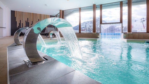 An indoor swimming pool with a modern water feature and large windows offering a view of snowy mountains. - Appartement Confortable Au Pied des Pistes | Wi-Fi Gratuit + Parking Disponible (Villarembert)