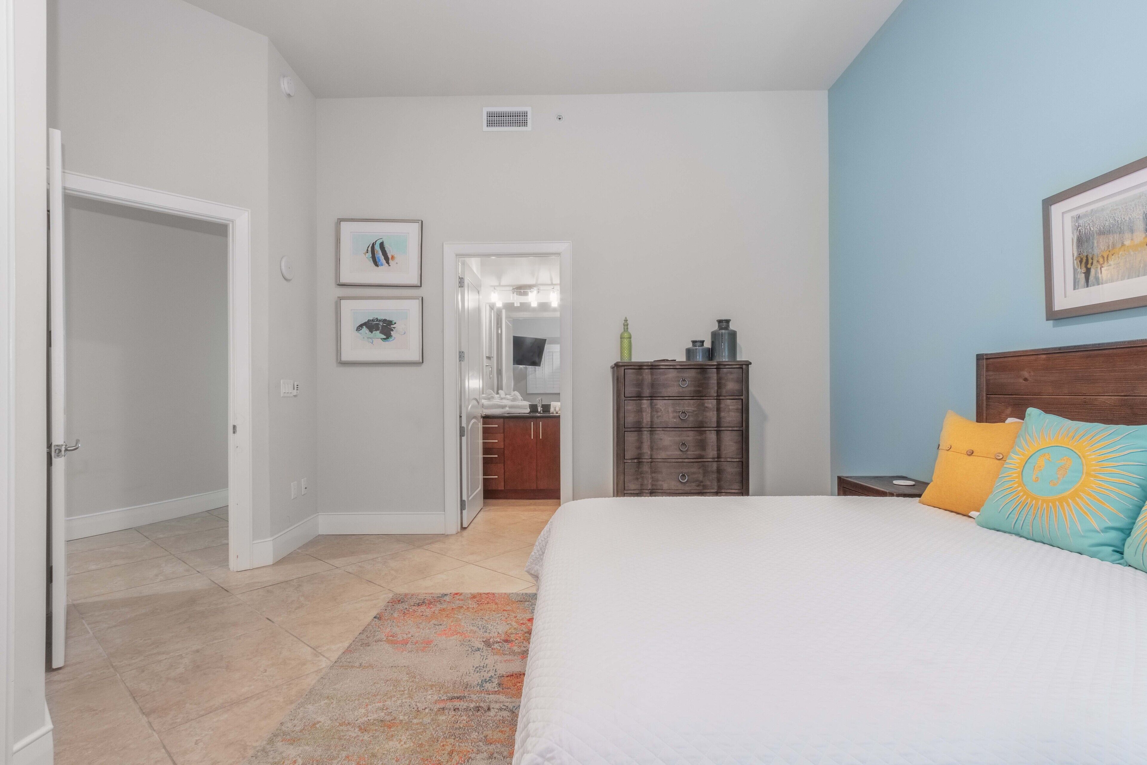 A bedroom with a bed, a nightstand, a dresser, a mirror, and a door leading to a bathroom.