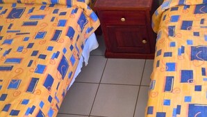 Zwei Einzelbetten mit passender gelb-blauer Bettwäsche, ein Nachttisch aus Holz mit einer Schublade und ein gefliester Boden.