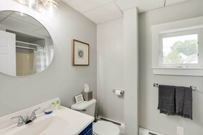 Une salle de bain avec un lavabo blanc, un miroir, un tableau encadré, un porte-serviettes et une fenêtre.