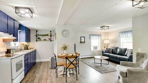Eine moderne Küche mit blauen Schränken, ein Esstisch mit Stühlen, ein Wohnzimmer mit Sofa und Sessel sowie eine Uhr an der Wand.