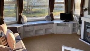 A cozy living room with a large window, a television, and a fireplace. - Immaculate 2-bed Static Caravan Near the Lake (Millom)