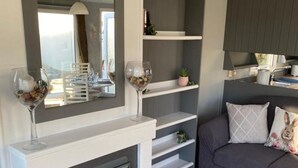 A modern living room with a fireplace, a sofa, and a dining area with a mirror and shelves. - Immaculate 2-bed Static Caravan Near the Lake (Millom)