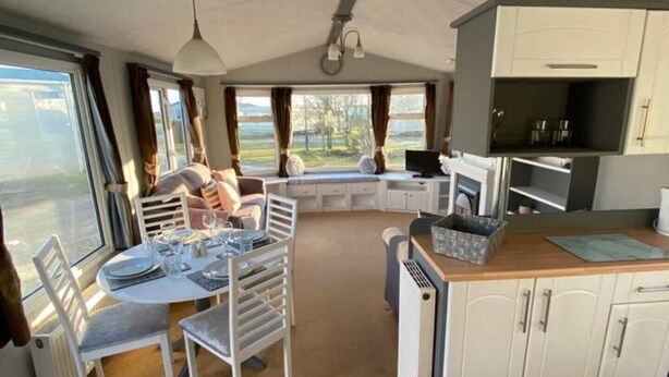 A compact kitchen with white cabinets, a dining area with a table set for four, and a large window offering a view of the outdoors. - Immaculate 2-bed Static Caravan Near the Lake (Millom)