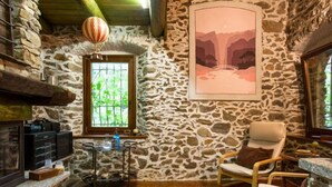 Um quarto rústico com paredes de pedra, teto de madeira, lareira, área de jantar com mesa e cadeiras, e uma grande janela com vista para o verde.