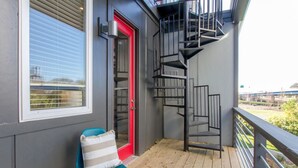 A wooden deck with a blue chair and a red door, a metal spiral staircase, and a view of a bridge in the background.
