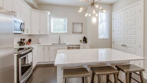 A modern kitchen with white cabinetry, a large island with bar stools, a stainless steel oven, and a unique chandelier.