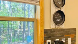 A kitchen with a window overlooking a forest, a coffee maker, and three decorative plates with sayings. - Romantic Splash Brand New 1 Bedroom Efficiency Cabin, near Broken Bow Lake (Broken Bow)