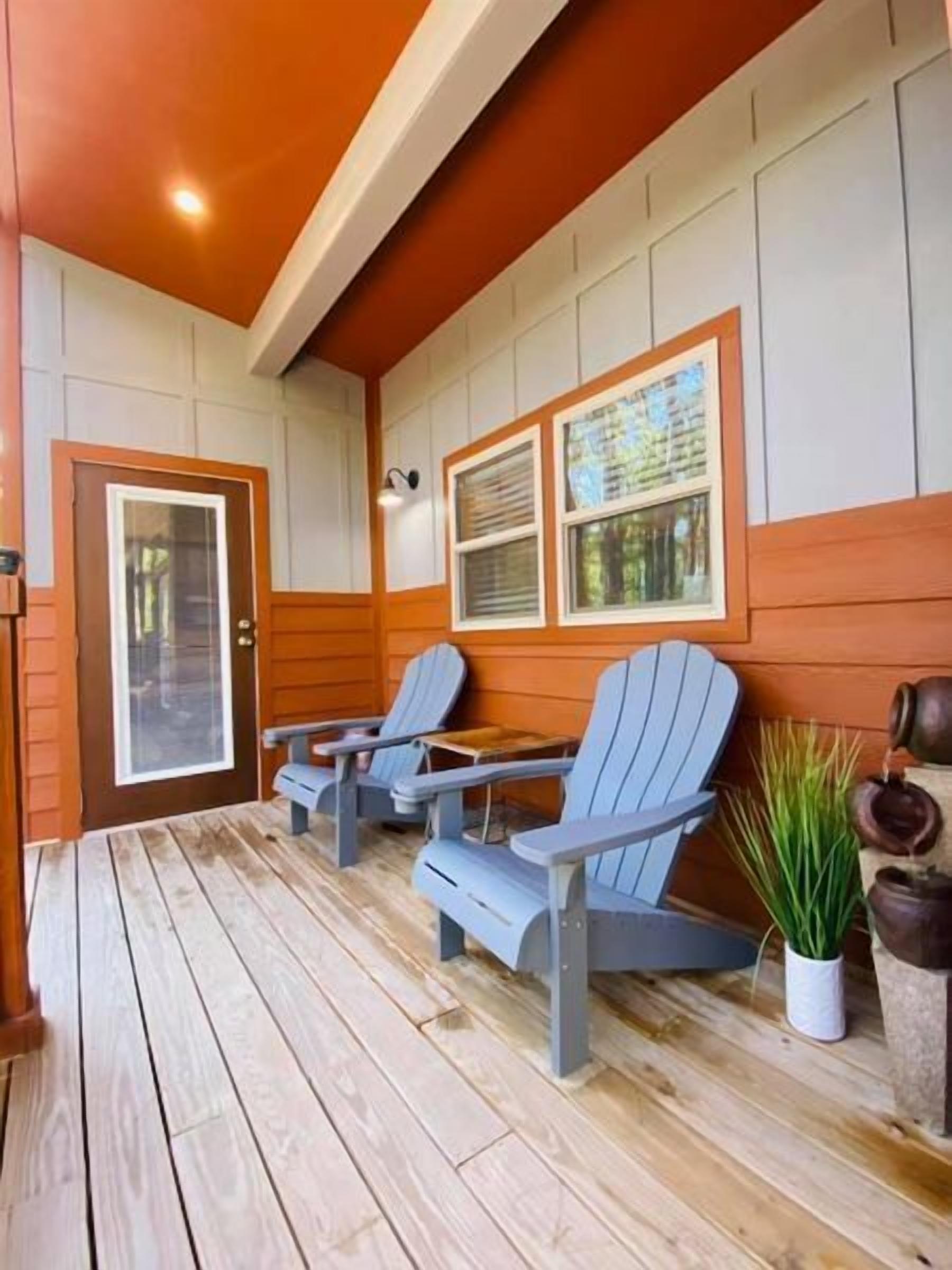 A covered porch with wooden flooring, two Adirondack chairs, a door with a glass panel, and a window with blinds.