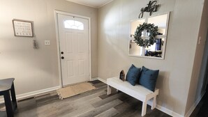 A hallway with a white door, a bench with blue cushions, a mirror with a wreath, and a sign with 'gather' on the wall. - The Flippin Fisherman  (Flippin)