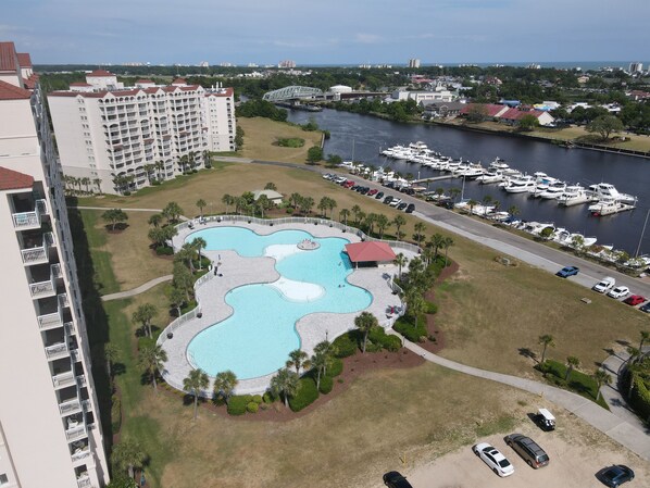 An aerial view of a resort with a large, irregularly shaped swimming pool surrounded by palm trees and a parking area.