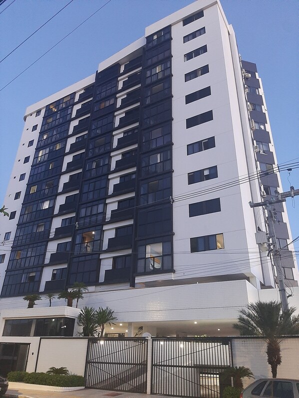 A multi-story building with a modern architectural design, featuring a combination of white and dark-colored panels, multiple balconies, and a gated entrance.