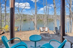 Un espace extérieur abrité, aménagé avec des chaises et une table, offrant une vue sur un lac bordé d’arbres et sur un quai en arrière-plan.