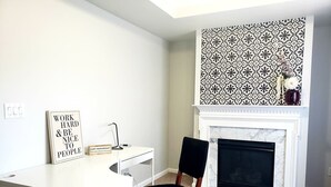 A room with a white desk, a black chair, a fireplace with a patterned wall above it, and motivational wall art.
