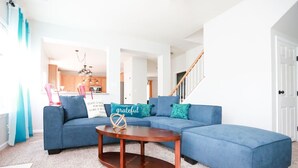 A modern living room with a blue sectional sofa, a wooden coffee table, and a kitchen in the background.
