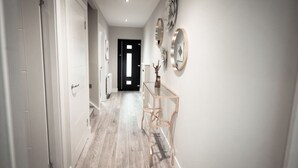 Un couloir doté d’un plancher en bois, de murs blancs et d’une porte au fond. On y trouve trois miroirs fixés au mur et une petite table sur laquelle est posé un vase.