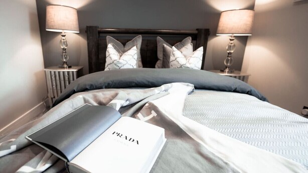 Un lit bien fait, sur lequel repose un livre intitulé « PRADA », deux lampes de chevet et une tête de lit agrémentée de coussins décoratifs.