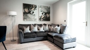 Un salon moderne avec un canapé d’angle gris, deux tableaux abstraits, une lampe sur pied et une porte.