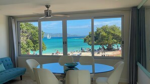 Quarto com uma grande janela que oferece vista para a praia e o oceano. Há uma mesa redonda com cadeiras e um ventilador de teto.