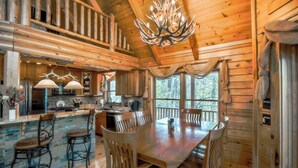 L’intérieur d’un chalet rustique est agrémenté d’une table à manger en bois, de tabourets de bar, d’une cuisine avec un mur arrière en pierre et d’une grande fenêtre offrant une vue sur la forêt.