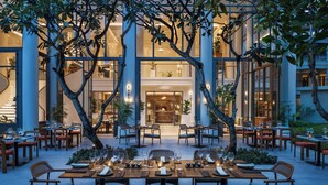 An outdoor dining area with tables and chairs, surrounded by trees and a building with large windows. - Centara Reserve Samui (Koh Samui)