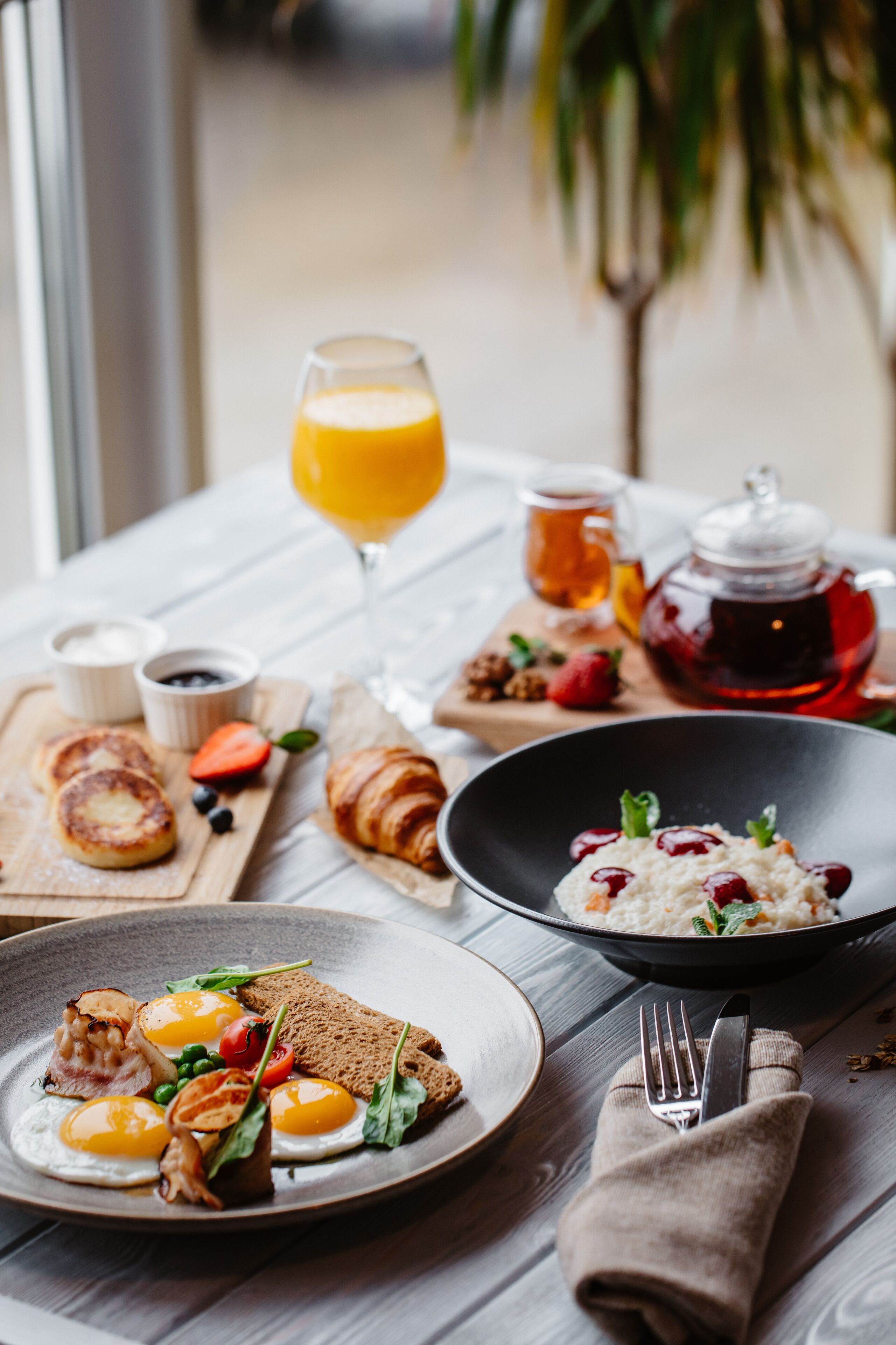 Un desayuno con diversos platos, entre ellos huevos, tostadas y un tazón de cereal con bayas. Hay un vaso de jugo de naranja y una tetera sobre la mesa.