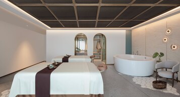 A modern spa room with two massage tables, a large freestanding bathtub, and a seating area with a chair and a small table.