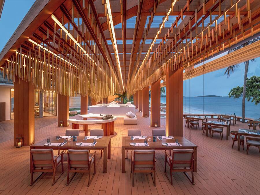 Un vaste espace extérieur aménagé pour les repas, avec des tables et des chaises en bois, un coin salon central et une vue sur l’océan.