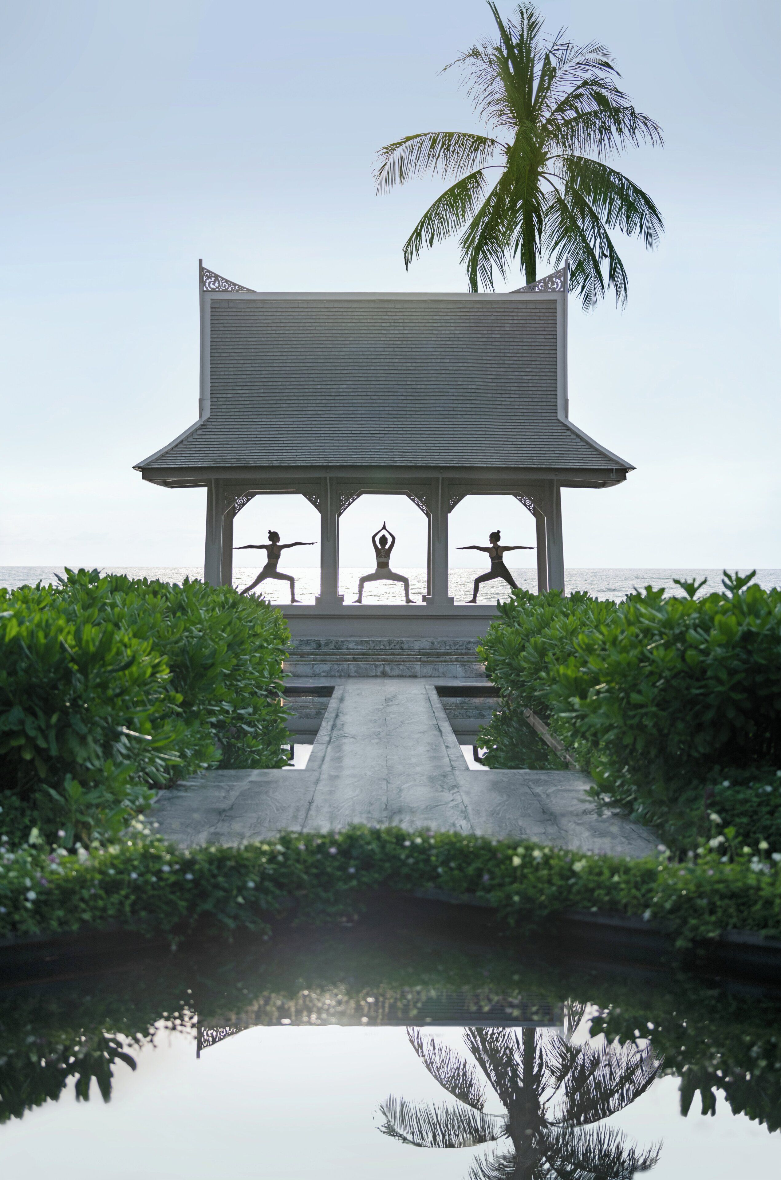 A gazebo with three silhouettes of people practicing yoga, a palm tree, and a body of water reflecting the scene.