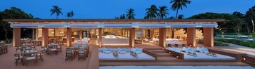 Un área de comedor al aire libre con mesas y sillas de madera, un espacio de descanso cubierto con cojines, y una piscina al fondo.