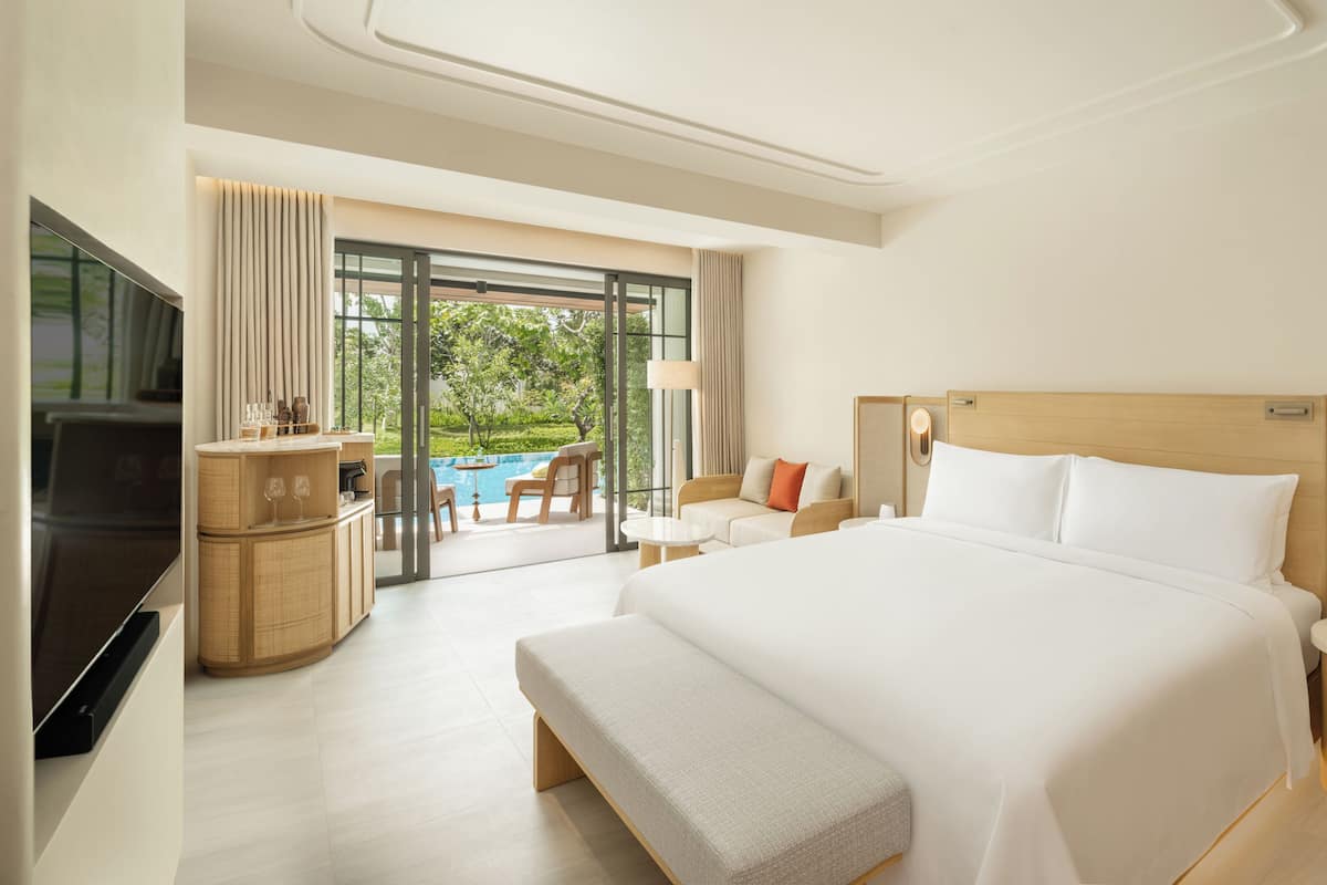 A hotel room with a large bed, a TV, a minibar, a seating area with a view of a pool, and a balcony.