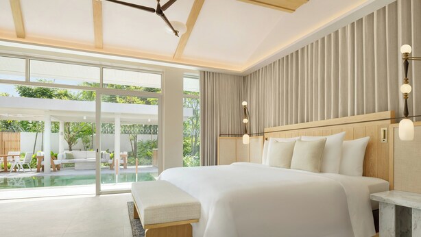 Habitación de hotel moderna con una cama grande, un banco, una mesita de noche y vistas a una zona de piscina exterior.