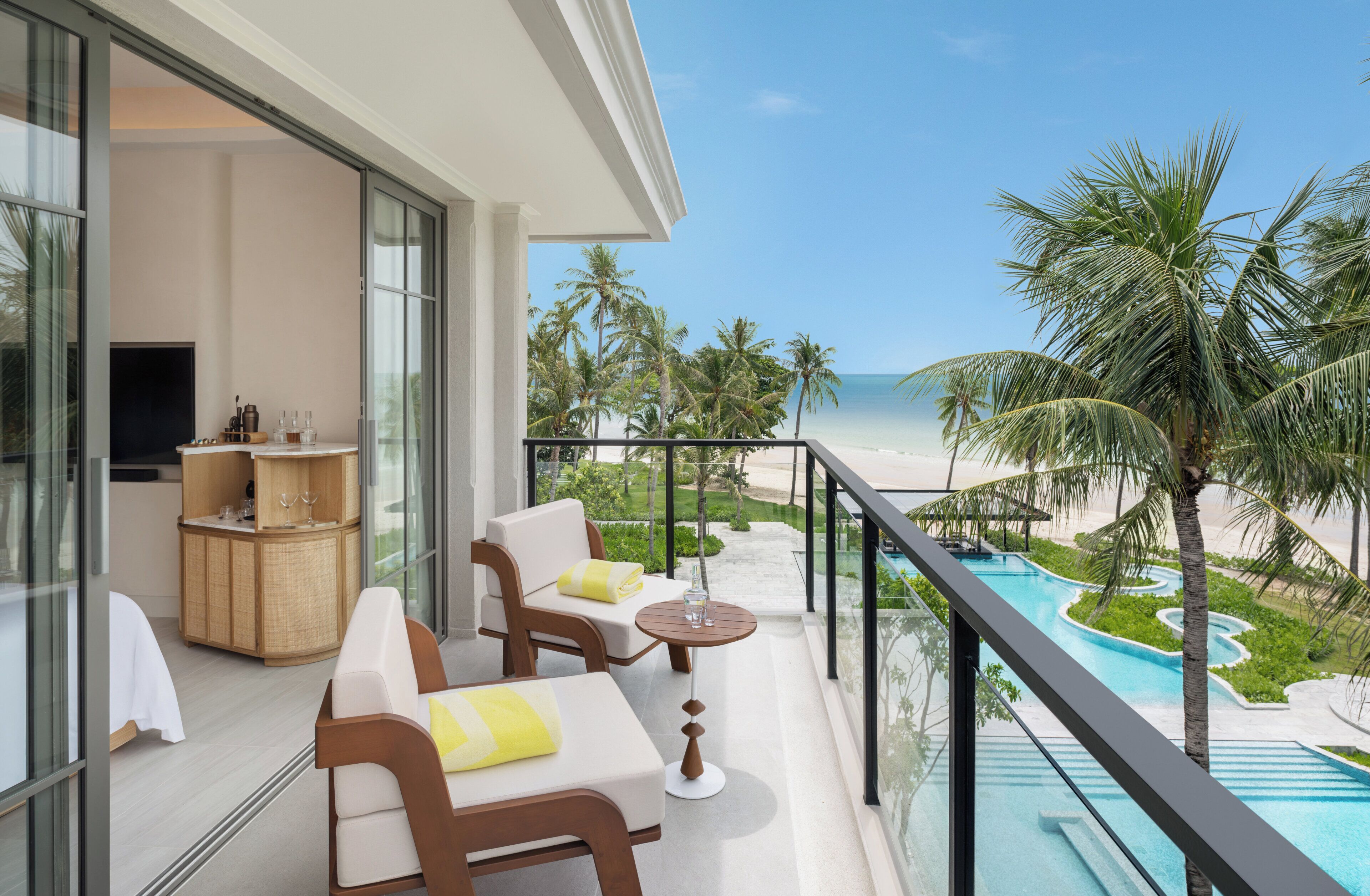 A balcony with a pool, white furniture, and a view of the beach and palm trees.