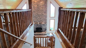 Una escalera de madera conduce a una sala de estar con chimenea de piedra, barandas de madera y un ventilador de techo.