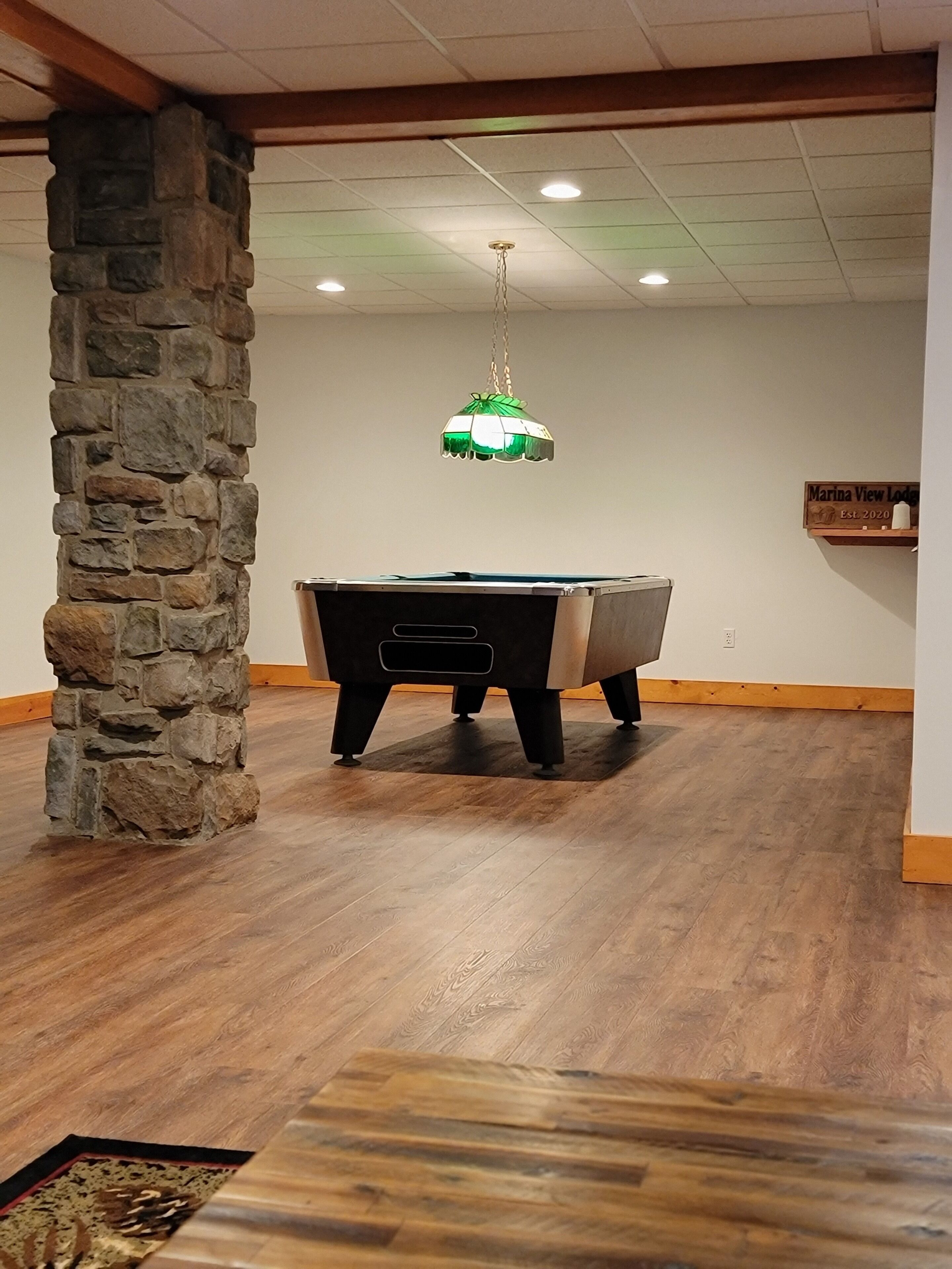 Una mesa de billar en una habitación con suelo de madera, un pilar de piedra y una lámpara verde colgante.