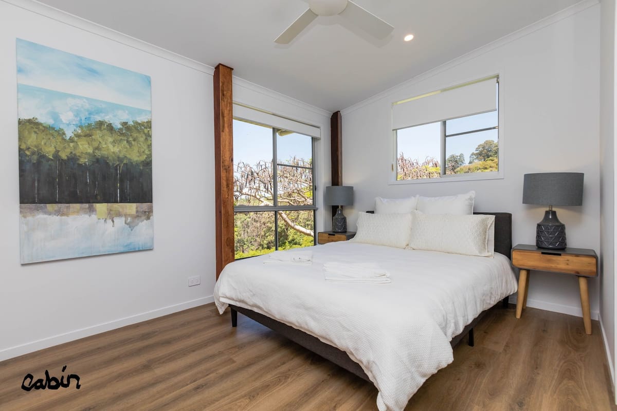 A bedroom with a large bed, two bedside tables, a ceiling fan, and a view of trees through the window.
