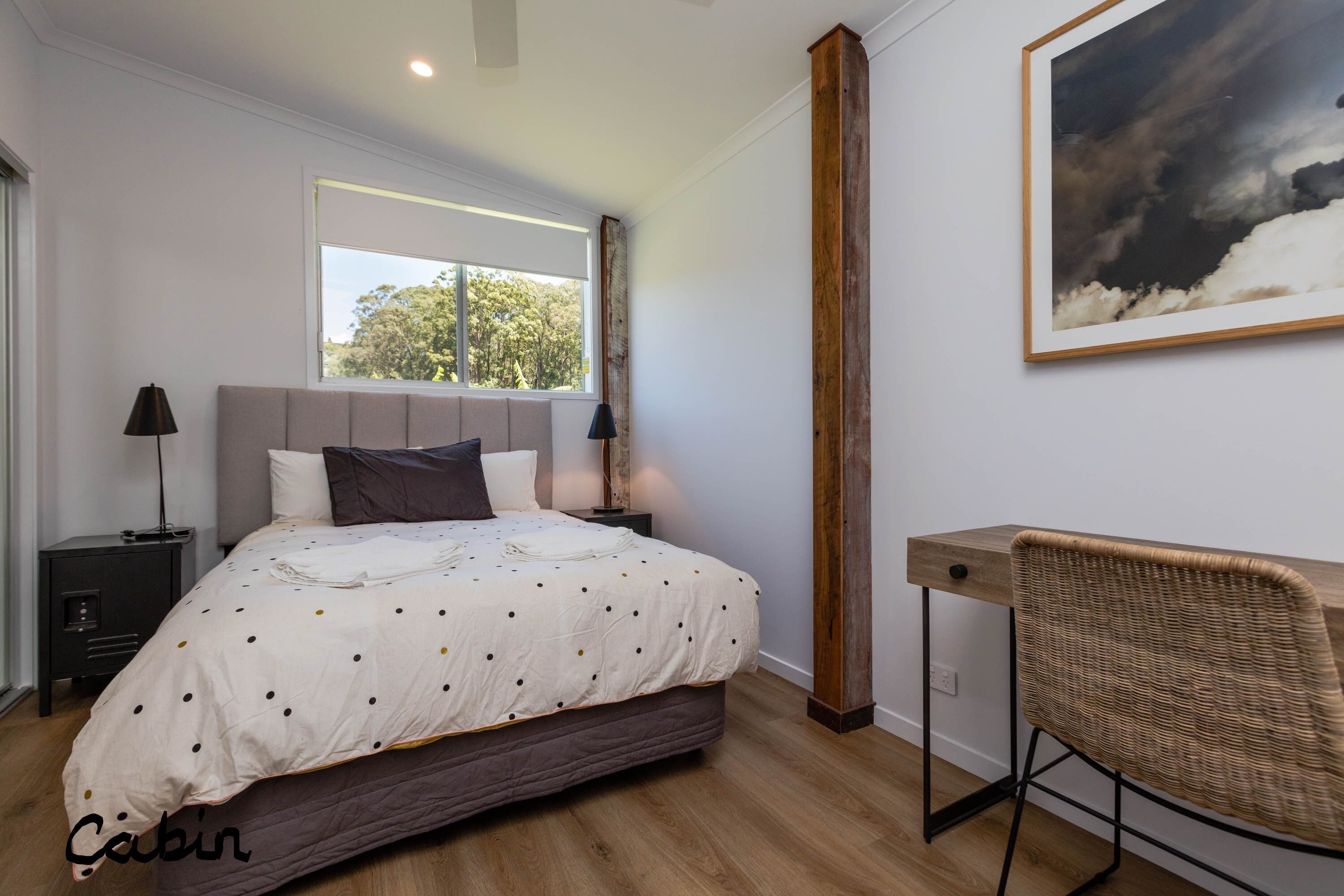 Un dormitorio con cama, mesitas de noche, un escritorio, una silla, una ventana y un cuadro en la pared.