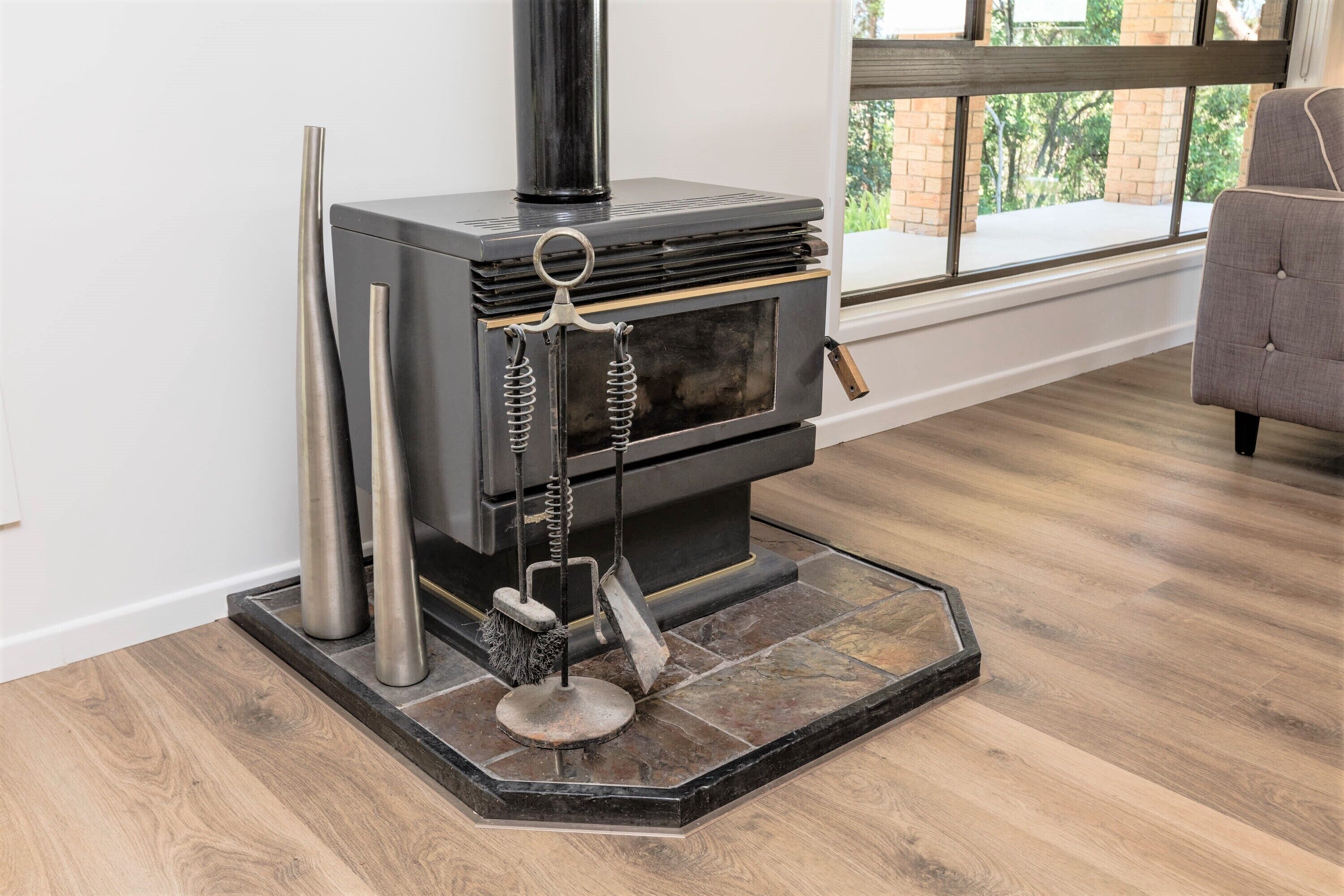 A modern wood-burning stove with a chimney, a brush, and a metal stand in a room with wooden flooring and a window.
