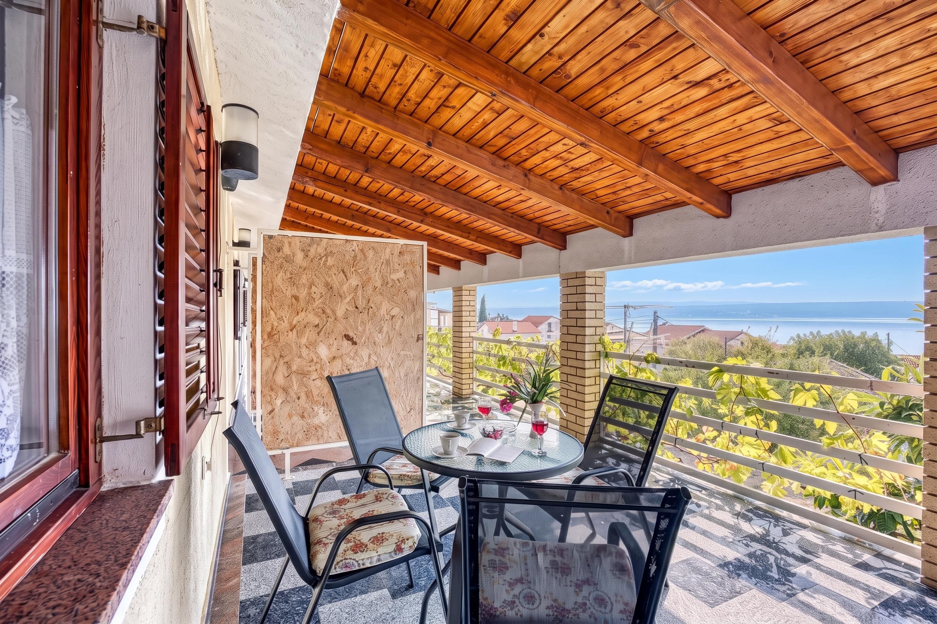 A balcony with a wooden ceiling, a glass table with chairs, and a view of a vineyard and sea.