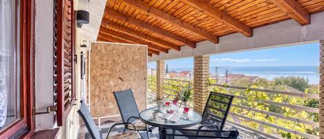 A balcony with a wooden ceiling, a glass table with chairs, and a view of a vineyard and sea.