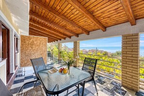 Un balcón con techo de madera, una mesa de vidrio con sillas, un jarrón con flores y vista al mar.