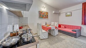 Een kleine keuken met een kookplaat, pannen en een spoelbak. Er is een eettafel met stoelen en een rode bank. Een schilderij en een lijst met een foto sieren de muren.