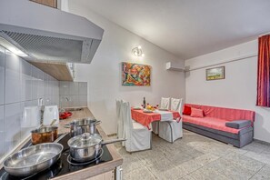 Cozinha compacta com fogão, panelas e pia. Há uma mesa de jantar com cadeiras e um sofá vermelho. Um quadro e uma fotografia emoldurada decoram as paredes.