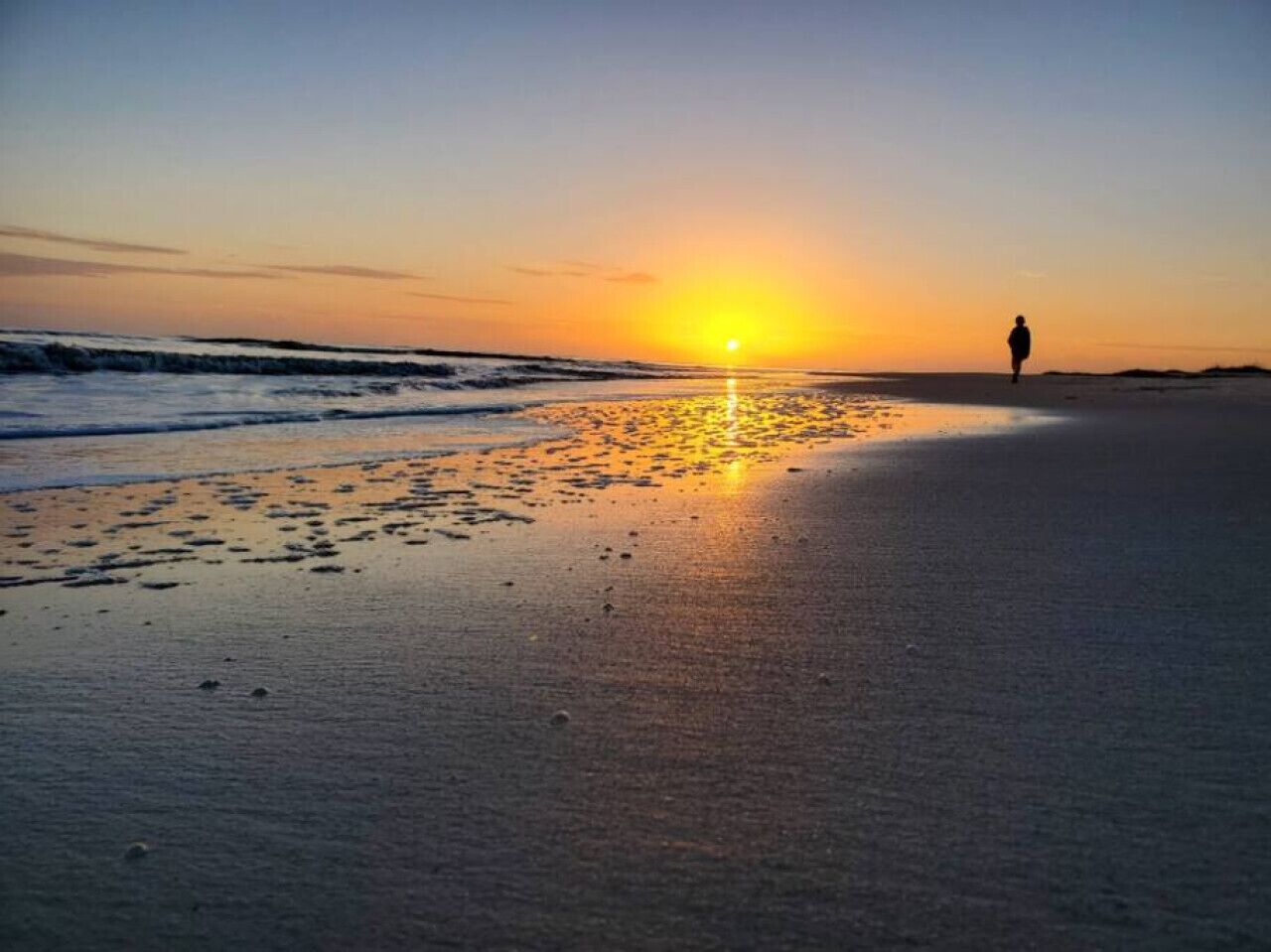 Ein Strand bei Sonnenuntergang mit Fußabdrücken, die zum Wasser führen, und einer Person, die vom Betrachter wegscheidet.