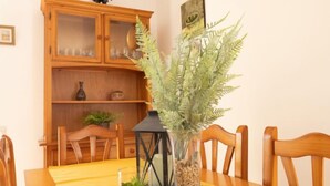 Una mesa de comedor con mantel amarillo, rodeada de sillas de madera. En el centro, un florero de vidrio con una planta y un farol. Detrás de la mesa, un aparador de madera con puertas de cristal y una repisa con adornos.