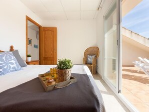 Een slaapkamer met een bed, een houten nachtkastje, een rieten stoel en een balkon met ligbedden.