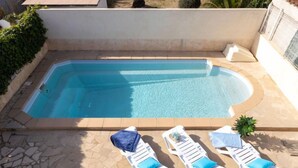 Una piscina rectangular con un borde de piedra, rodeada de sillones blancos con cojines azules.
