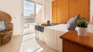Un dormitorio con cama, armario de madera, silla de mimbre, una mesita con una planta y puerta corredera de cristal que da a un espacio exterior.