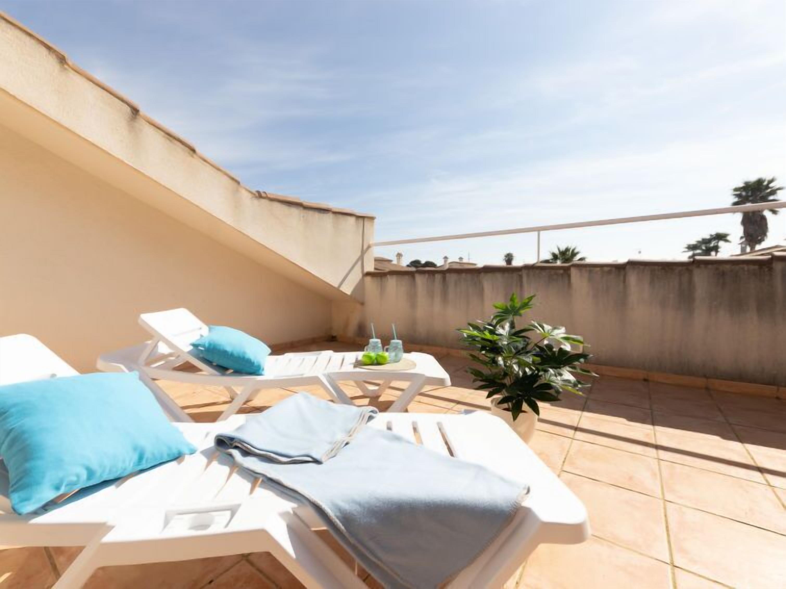 Une terrasse sur le toit, avec des chaises longues blanches, des coussins bleus, une petite table avec des boissons et une plante en pot.